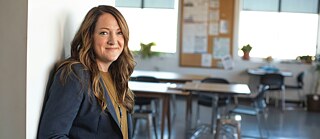 Person mit langen Haaren in einem Business-Outfit lehnt an einer Wand in einem hellen Raum mit mehreren Tischen und Stühlen. Im Hintergrund sind große Fenster, eine Pinnwand mit Papieren und einige Pflanzen zu sehen.