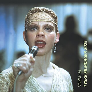 Person in elegantem, goldfarbenem Outfit hält ein Mikrofon auf einer Bühne, im Hintergrund unscharfe Musiker und Instrumente, darunter eine Gitarre. 