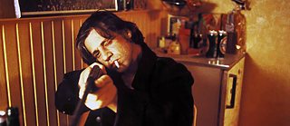 Person in dunkler Kleidung zielt mit einer Schrotflinte in einem holzvertäfelten Raum. 