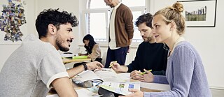 A group of people sits at a table in a bright room, working together. Pens, brochures, and study materials are on the table, with windows and wall decorations in the background.
