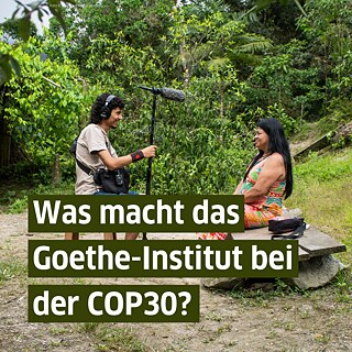 Eine Interviewszene in einem Wald, in der eine auf einer Bank sitzende indigene Frau und ein ihr gegenübersitzender Interviewer mit Kopfhörern und Stabmikrofon zu sehen sind.