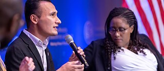 Two people on a stage. A man speaks into a microphone