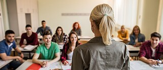Eine Lehrerin steht vorne im Klassenraum und spricht zu einer Gruppe junger Erwachsener, die aufmerksam zuhören. Die Lernenden sitzen an Tischen mit Heften und Papieren vor sich und lächeln. Der Raum ist hell und freundlich mit großen Fenstern auf der rechten Seite. 