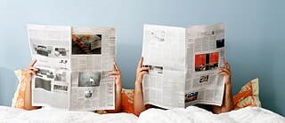 Zwei Personen liegen im Bett und lesen große Zeitungen, Gesichter sind vollständig verdeckt.