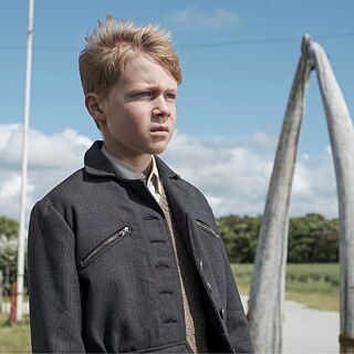 A close-up of a boy with light hair and a dark jacket looking toward the right edge of the picture, with a meadow, trees, and blue sky in the background.