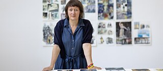 Barbara Yelin steht hinter einem Schreibtisch; hinter ihr eine Wand mit verschiedenen Illustrationen der Künstlerin.