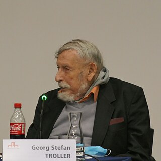 Das Bild zeigt Georg Stefan Troller, der an einem Tisch sitzt, vermutlich bei einer Veranstaltung oder einem Vortrag. Er trägt ein dunkles Sakko mit einer bordeauxfarbenen Einstecktasche, darunter ein orangefarbenes Hemd und einen hellgrauen Pullover. Vor ihm stehen mehrere Glasgefäße: eine Karaffe, ein Trinkglas und eine weitere Karaffe auf der linken Seite. Links im Vordergrund befindet sich eine Flasche Coca-Cola mit rotem Schraubverschluss. Direkt vor Troller ist ein Mikrofon mit einer kleinen grünen Leuchtanzeige zu sehen. 