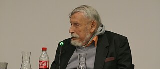 Das Bild zeigt Georg Stefan Troller, der an einem Tisch sitzt, vermutlich bei einer Veranstaltung oder einem Vortrag. Er trägt ein dunkles Sakko mit einer bordeauxfarbenen Einstecktasche, darunter ein orangefarbenes Hemd und einen hellgrauen Pullover. Vor ihm stehen mehrere Glasgefäße: eine Karaffe, ein Trinkglas und eine weitere Karaffe auf der linken Seite. Links im Vordergrund befindet sich eine Flasche Coca-Cola mit rotem Schraubverschluss. Direkt vor Troller ist ein Mikrofon mit einer kleinen grünen Leuchtanzeige zu sehen. 