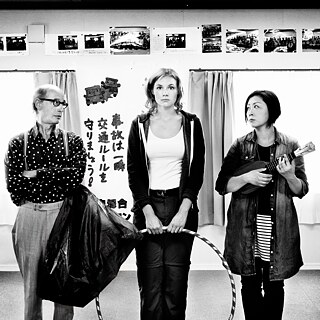 Das schwarz-weiß Foto zeigt drei Personen. Eine Person in der Mitte schaut in die Kamera und die anderen beiden links und rechts blicken zur Person in der Mitte. Eine Person hält einen Reifen und eine Person hält ein Instrument.