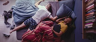 Das Bild zeigt von oben fotografiert zwei Personen auf dem Bett liegend, eine Person hat den Laptop in der Hand.