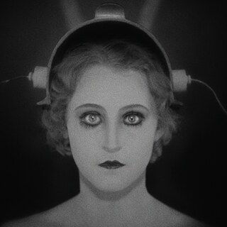 Das Foto ist ein schwarz-weiß Bild und zeigt eine Person stark geschminkt in die Kamera blickend mit einem verkabeltem Gerät auf dem Kopf.