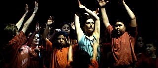 Na foto, há várias pessoas a levantar os braços. Elas estão bem iluminadas e o fundo é bastante escuro.