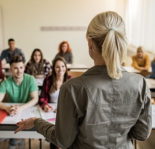 	Rückansicht auf eine Lehrerin, die zu Schülern der Sekundarstufe im Klassenraum spricht.