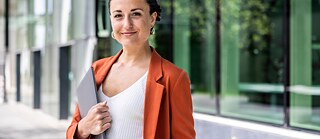 Person wearing an orange blazer and white top holding a laptop in front of a modern glass building.