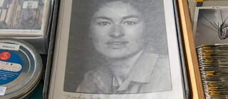 „Archivaufnahme mit verschiedenen Materialien: links eine Filmrolle mit blauem Etikett, in der Mitte ein großformatiges Schwarz-Weiß-Foto in einer Mappe, rechts ein Stapel von Dokumenten und eine Fotografie mit einer modernen Skulptur. Die Szene zeigt historische und künstlerische Archivbestände.“