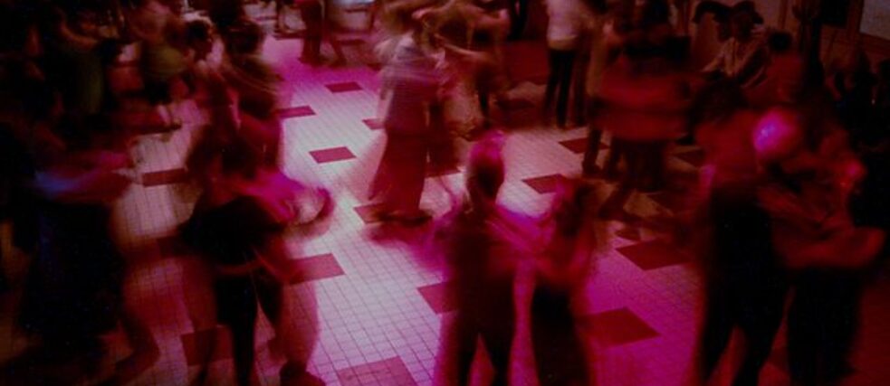 Es ist ein Ballsaal in rosa Licht zu sehen, in dem viele Paare tanzen