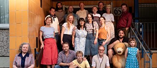 Foto grupal de varias personas que están de pie y sentadas en los escalones de un edificio residencial. Las personas visten ropa cotidiana de verano, y a la derecha hay un gran oso de peluche marrón claro junto a una niña. La fachada del edificio está revestida con azulejos amarillos, y la escena recuerda a una foto comunitaria o familiar.