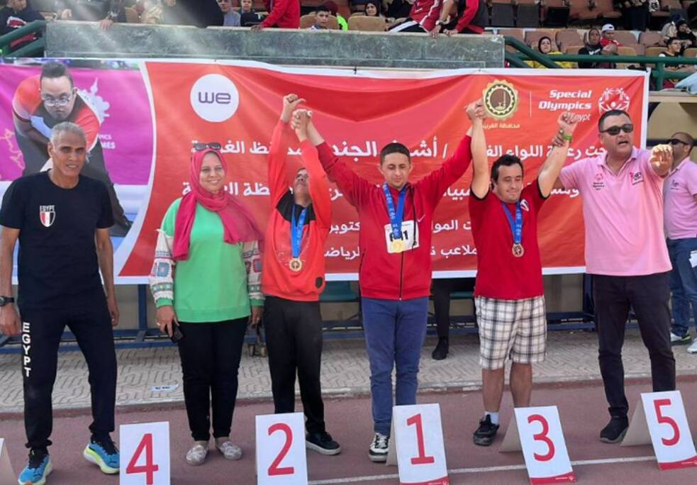 Drei Kinder stehen in der Mitte, links zwei Erwachsene, eine Frau und ein Mann, rechts, ein Mann. Sie haben fröhliche Gesichtsausdrücke und haben ihre Arme hoch erhoben. Die Kinder tragen alle Medaillen um den Hals.