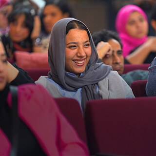 فتاة تجلس بين الحضور، تبتسم وتتابع الحديث باهتمام.