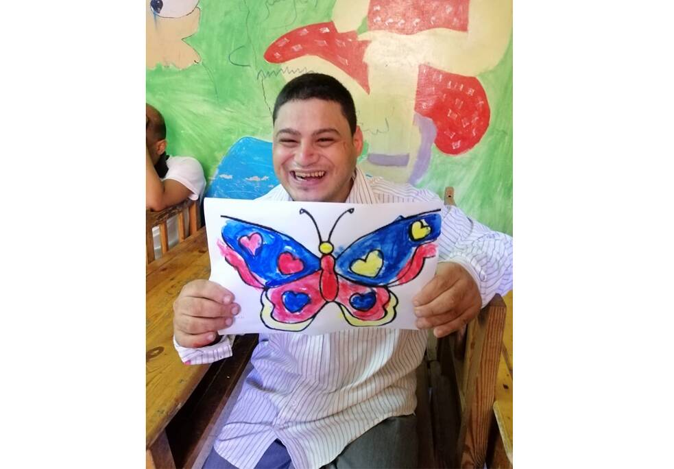 Ein Junge sitzt schräg an einem Tisch und schaut in die Kamera. Er hält einen selbstgebastelten, großen, blau-gelb-roten Schmetterling in die Kamera.