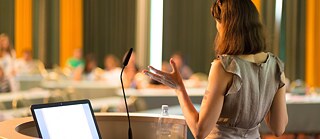 Das Bild zeigt eine Person am Rednerpult, die vor einem Publikum spricht. Auf dem Pult stehen ein Mikrofon, ein Laptop und eine Wasserflasche. Die Szene erinnert an eine Konferenz oder einen Redewettbewerb.