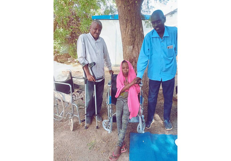 Ein Mädchen sitzt im Rollstuhl. Links von ihr steht ein Mann an Krücken. Rechts von ihr steht ein weiterer Mann, reicht ihr die Hand und lächelt in die Kamera.