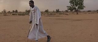Ein Mann läuft mit Krücken in einer sandigen Landschaft. Er trägt eine weißem Dschabbab (traditionelle Kleidung).  
