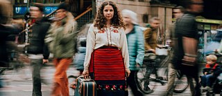 Eine Person steht in traditioneller Kleidung mit bestickter weißer Bluse und rot-schwarzem Rock mitten auf einer belebten Stadtstraße. In der Hand hält sie einen Koffer. Um sie herum bewegen sich viele Menschen und Fahrräder, die durch lange Bewegungsunschärfen verschwommen wirken, wodurch die stehende Person deutlich hervorsticht.