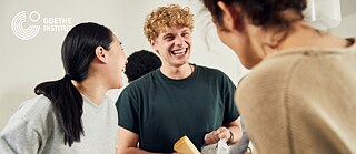 幾位人士在明亮的室內空間中互相交談，其中一人手上拿著杯子和紙張。背景中可見歌德學院的標誌。