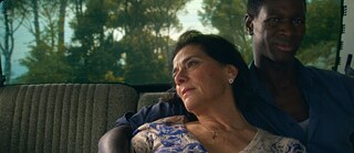 A couple sits in the back seat of a car. The man has his arm around the woman. The man is a young, Black man, and the woman is older and fair skinned..