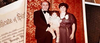 صورة عائلية قديمة. الأب، مرتدياً بدلة سوداء، يبتسم وهو يحمل ابنته الصغيرة بين ذراعيه. الأم، مرتديةً فستاناً طويلاً بلون خمري مزيناً بزهور بيضاء على الجانب الأيمن من صدرها، تبتسم هي الأخرى.