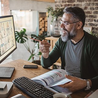 Una persona está sentada en un escritorio participando en una videoconferencia. En la pantalla del ordenador se ven varios participantes en una reunión en línea. La persona sostiene un bolígrafo y parece señalar la pantalla o explicar algo. El espacio de trabajo está organizado con plantas, documentos y un monitor moderno. Al fondo se observa una habitación con luz natural cálida y una pared de ladrillos a la vista.