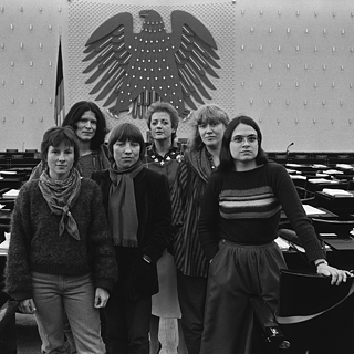 Un grupo de mujeres posa en una amplia sala oficial de sesiones, reconocible por su arquitectura característica y el emblemático águila federal en la pared, lo que indica un entorno político. Las personas se encuentran en el centro del espacio, rodeadas de asientos y mesas vacías que evocan un ambiente parlamentario.