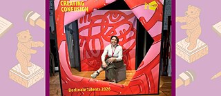 سيدة مبتسمة ترتدي سترة بيضاء تجلس وساقاها متقاطعتان أمام خلفية حمراء.