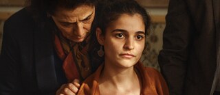 تنظر شابة إلى الأمام مباشرةً بتعبير قلق. امرأة مسنة تضع يدها اليمنى على كتفها وتهمس بشيء في أذنها.