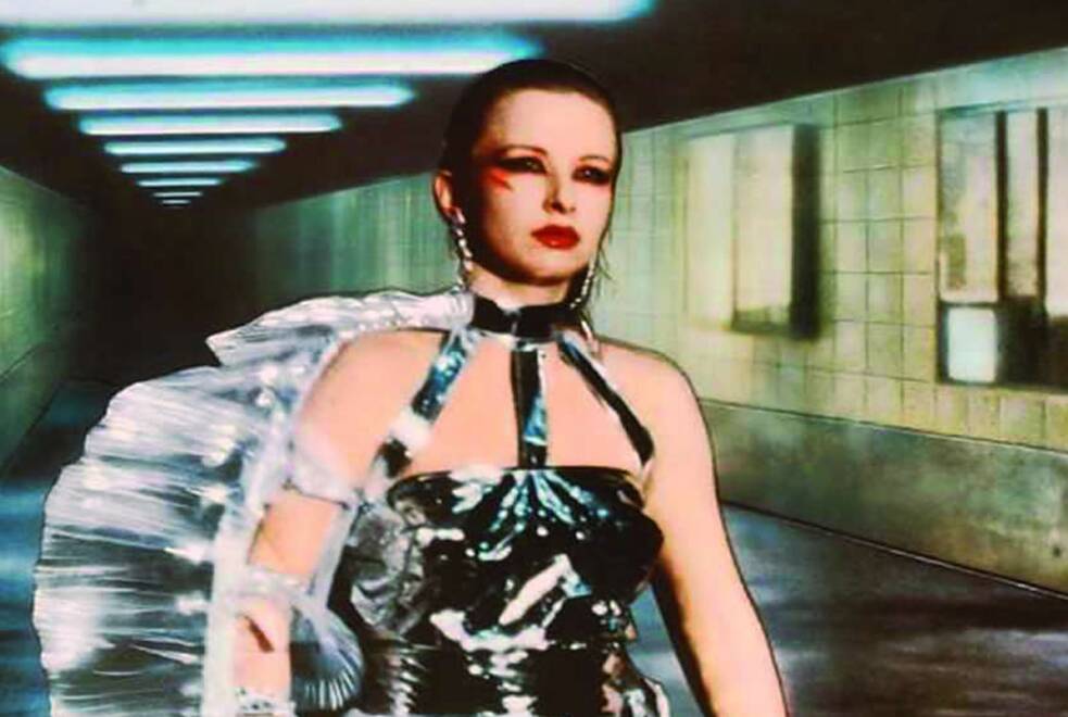 Standbild aus dem Film Bildnis einer Trinkerin. Eine Person mit kurzen Haaren geht durch einen langen, beleuchteten Tunnel mit gefliesten Wänden. Sie trägt ein glänzendes schwarzes Kleid mit Halsband, lange Ohrringe und auffälliges Augen-Make-up sowie roten Lippenstift; über einer Schulter liegt ein großer, transparenter Fächer. An der Decke verläuft eine Reihe rechteckiger Leuchten, der Hintergrund ist leicht unscharf. 