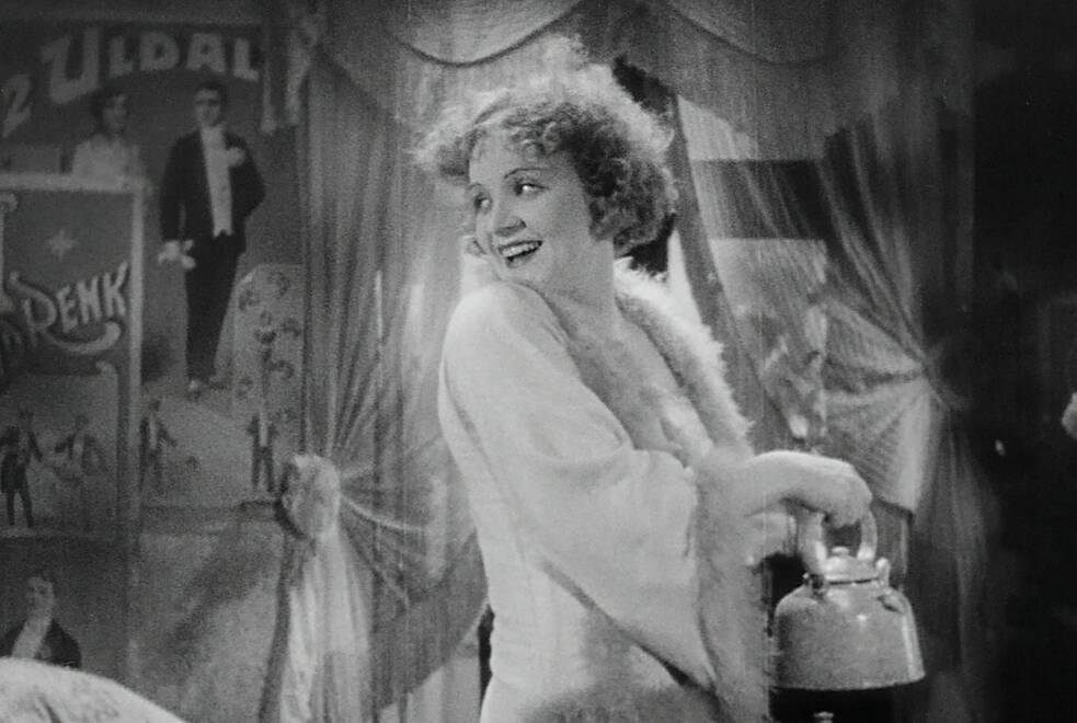 Standbild aus dem Film Der blaue Engel mit der Schauspielerin Marlene Dietrich: Eine Frau mit kurzen, lockigen Haaren steht seitlich und blickt nach links. Sie trägt ein helles Kleidungsstück mit pelzartigem Besatz und hält einen Metallkessel auf einer kleinen Herdplatte; in der anderen Hand hält sie ein dünnes Stoffstück. Im Hintergrund sind Vorhänge und mehrere Wandplakate mit abgebildeten Personen und Schriftzügen zu sehen.