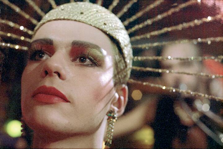 Standbild aus dem Film Coming Out. Nahaufnahme des Gesichts einer Person mit einer glitzernden Kopfbedeckung, aus der dünne, goldfarbene Stäbe strahlenförmig nach außen ragen. Die Person trägt rotes und goldfarbenes Augen-Make-up, roten Lippenstift und lange, hängende Ohrringe. Der Kopf ist leicht nach oben geneigt, im unscharfen Hintergrund sind Lichtpunkte und Formen zu sehen.