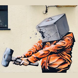 Das Streetart-Werk des palästinensischen Künstlers Taqi Spateen zeigt eine Figur in einer leuchtend orangefarbenen Jacke, deren Kopf als aufbrechender Betonblock dargestellt ist, während sie mit einem Hammer darauf einschlägt. Die kraftvolle, dynamische Szene wirkt wie ein Symbol für Widerstand und den Versuch, starre Strukturen oder innere Barrieren zu durchbrechen