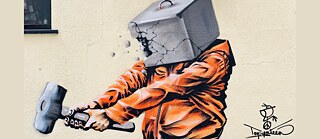 Das Streetart-Werk des palästinensischen Künstlers Taqi Spateen zeigt eine Figur in einer leuchtend orangefarbenen Jacke, deren Kopf als aufbrechender Betonblock dargestellt ist, während sie mit einem Hammer darauf einschlägt. Die kraftvolle, dynamische Szene wirkt wie ein Symbol für Widerstand und den Versuch, starre Strukturen oder innere Barrieren zu durchbrechen