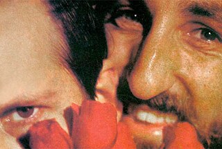 Standbild aus dem Film Taxi zum Klo. Das Bild zeigt eine Nahaufnahme von zwei Personen. Im Vordergrund ist das Gesicht der linken Person teilweise sichtbar, besonders das linke Auge, vor dem sich rote Blütenblätter befinden. Die rechte Person ist sehr nah neben der linken, man sieht Nase, Mund und Augen, der Mund ist leicht geöffnet und die Zähne sind sichtbar.