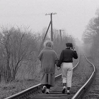 Foto en blanco y negro de dos mujeres vistas desde atrás, caminando juntas por una vía de tren abandonada. Avanzan muy cerca una de la otra, tomadas de la mano, hacia un paisaje con niebla y árboles sin hojas.