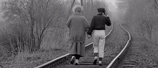 Foto en blanco y negro de dos mujeres vistas desde atrás, caminando juntas por una vía de tren abandonada. Avanzan muy cerca una de la otra, tomadas de la mano, hacia un paisaje con niebla y árboles sin hojas.