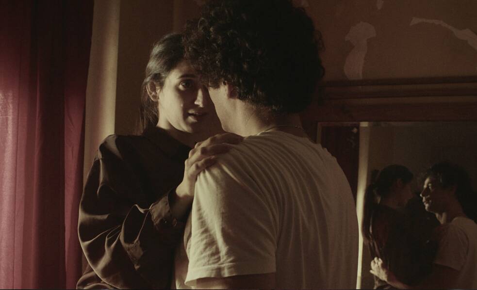 لقطة مقرّبة لامرأة ورجل يقفان متقاربين في غرفة، تضع المرأة يدها على كتفه. وخلفهما مرآة تعكس صورتهما.