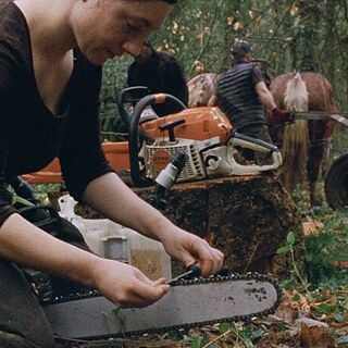 Eine Person arbeitet im Wald an einer Kettensäge und bereitet die Kette sorgfältig vor. Im Hintergrund ziehen zwei Menschen mit Hilfe eines Pferdes Holz über den Waldboden
