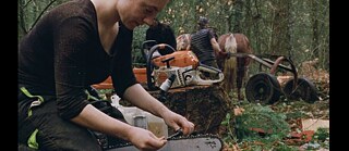 Eine Person arbeitet im Wald an einer Kettensäge und bereitet die Kette sorgfältig vor. Im Hintergrund ziehen zwei Menschen mit Hilfe eines Pferdes Holz über den Waldboden.