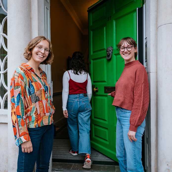 On the left and right, two young white women smile at the camera and welcome you into Goethe-Institut Irland to learn German courses. The green door at 37 Merrion Square is open, and another young woman can be seen from behind going in.