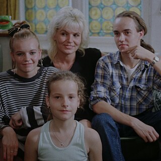 Eine Familiengruppe von vier Personen sitzt eng beieinander in einem Wohnzimmer. Hinter ihnen befindet sich ein Fenster mit farbigen, kreisförmigen Mustern. Links steht ein Fernsehgerät, rechts ein Tisch mit einem Stoffbezug und einem Radio. 