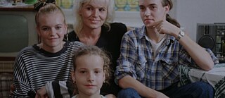 Un grupo familiar de cuatro personas está sentado junta en una sala de estar. Detrás de ellas hay una ventana con patrones circulares de colores. A la izquierda se ve un televisor y a la derecha una mesa con un mantel y una radio. 