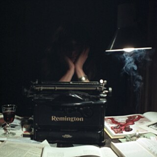 Una habitación oscura con un escritorio sobre el que hay una máquina de escribir negra de la marca Remington. El escritorio está cubierto de libros abiertos, papeles y un dibujo con líneas rojas. A la izquierda hay una copa con una bebida oscura y un objeto de vidrio azul. A la derecha cuelga una lámpara, bajo la cual se eleva humo. Detrás de la máquina de escribir está sentada una persona, con la cabeza apoyada en las manos.